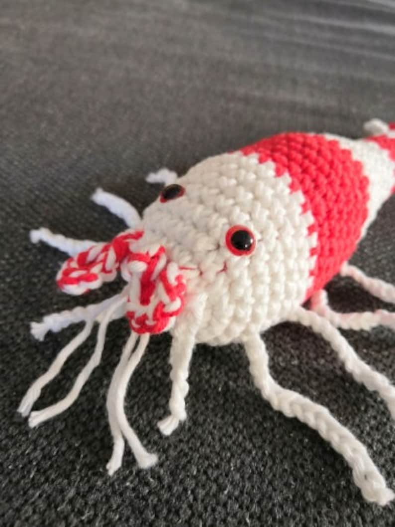 K&ouml;nnte beinhalten: Ein geh&auml;keltes Garnelenspielzeug in Wei&szlig; und Rot. Die Garnele hat einen segmentierten K&ouml;rper mit roten Streifen und kleinen schwarzen Augen. Sie hat wei&szlig;e Antennen und Beine. Das Spielzeug ist aus Garn gefertigt und soll wie eine Garnele aussehen.