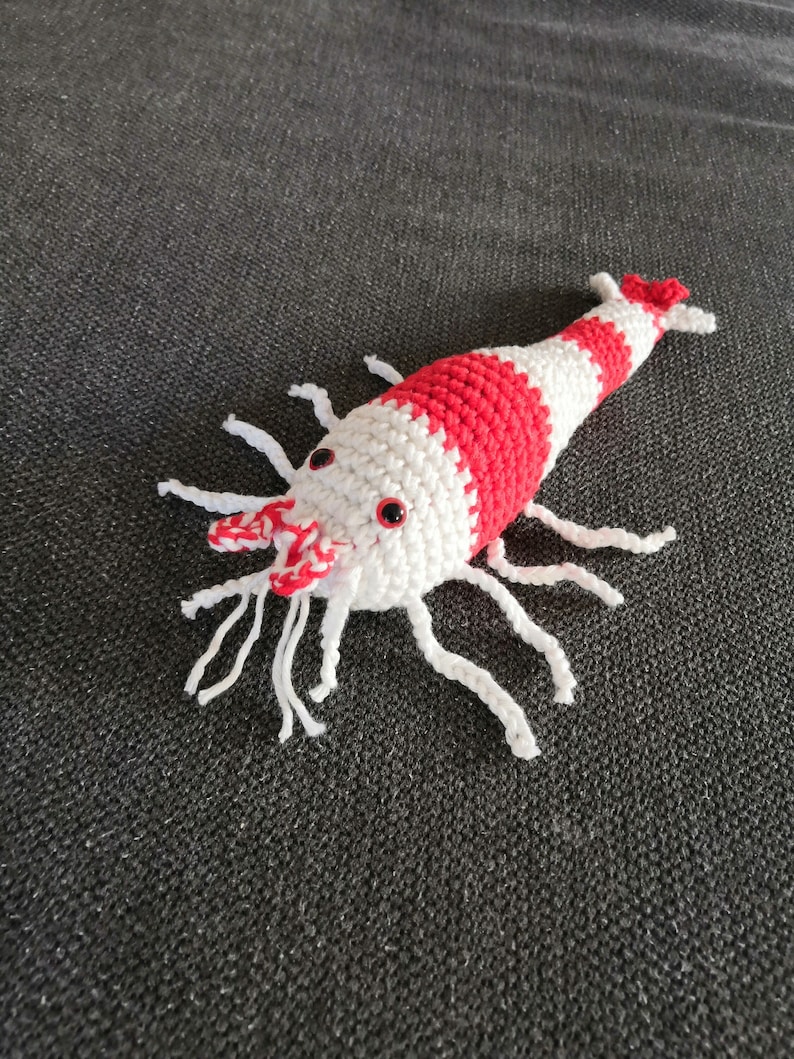 K&ouml;nnte beinhalten: Eine handgefertigte H&auml;kelgarnele. Die Garnele hat einen wei&szlig;en K&ouml;rper mit roten Streifen und wei&szlig;en Fadenantennen. Die Augen sind klein und rot. Dieses weiche Spielzeug ist ein einzigartiges Geschenk oder ein lustiges Dekorationsobjekt, etwa 15 cm lang.