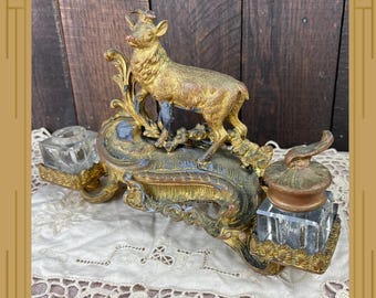 May include: A gold-toned metal inkstand featuring a central deer figurine standing on an ornate, textured base. The stand includes two clear glass inkwells, one with a matching metal lid, positioned on either side of the central decorative element.