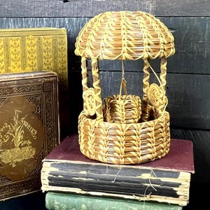 May include: A miniature woven basket well with a domed roof, suspended bucket, and decorative accents. The well is made of light brown woven material and sits atop a stack of antique books with gold lettering and ornate covers.