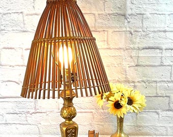 May include: A table lamp with a gold-toned metal base and a conical shade constructed from vertical brown wooden slats. The lamp features a central light bulb, a decorative pedestal stand with embossed detailing, and a square base with four feet.