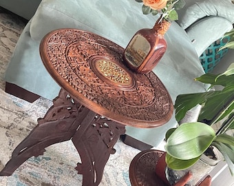 May include: A round, dark brown wooden side table with intricate floral carvings and a central brass inlay. The table features a folding base with cutout details. A glass bottle with a peach-colored flower and a green plant are placed on top.