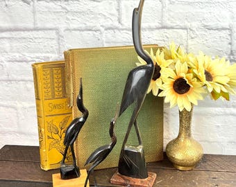 May include: Three dark, polished horn bird sculptures standing on wooden bases, arranged in front of a yellow book titled Swiss, a green book, and a brass vase filled with yellow sunflowers against a white brick wall.