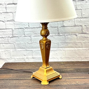 Puede incluir: Una lámpara de mesa dorada con una pantalla de color crema. La lámpara tiene una base decorativa con detalles ornamentales y una base cuadrada. Está sobre una superficie de madera contra una pared de ladrillos blancos.