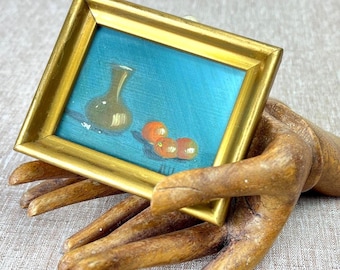 Peut inclure: Une petite peinture rectangulaire dans un cadre doré représentant une nature morte avec un vase clair et trois fruits orange sur un fond bleu sarcelle vif, présentée sur un modèle de main articulé en bois.