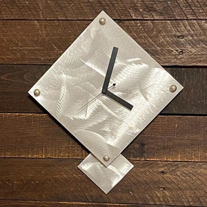 Pode incluir: Um relógio de parede moderno em forma de diamante, com acabamento em metal escovado. O relógio tem ponteiros pretos e está montado em um fundo de madeira. O diâmetro do relógio é de cerca de 30 cm e possui rebites prateados em cada canto.