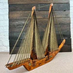 Peut inclure: Maquette de voilier en bois avec deux voiles en fil beige et noir. Le bateau a une coque et un mât en bois brun foncé, avec une base en bois brun clair. Les voiles sont faites de fil tendu.