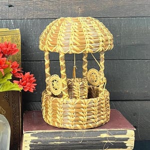 May include: A woven straw wishing well ornament with a domed roof, positioned atop a stack of two vintage books. Beside the books, a clear glass bottle holds a bouquet of artificial red flowers with green leaves, creating a decorative display.