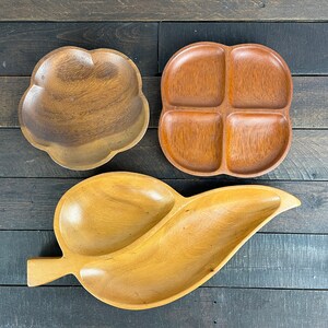 Pode incluir: Três travessas de madeira para servir em várias formas. Uma tem formato de flor, outra tem quatro compartimentos e a terceira tem formato de folha. Os tons de madeira variam de castanho claro a médio, sobre um fundo de madeira escura.