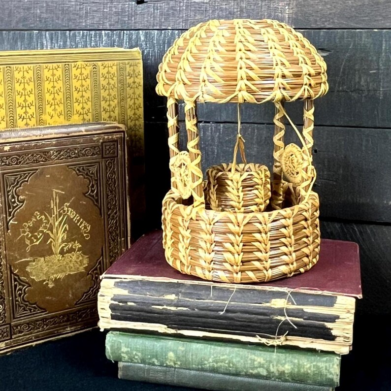 May include: A miniature woven basket well with a domed roof, suspended bucket, and decorative accents. The well is made of light brown woven material and sits atop a stack of antique books with gold lettering and ornate covers.