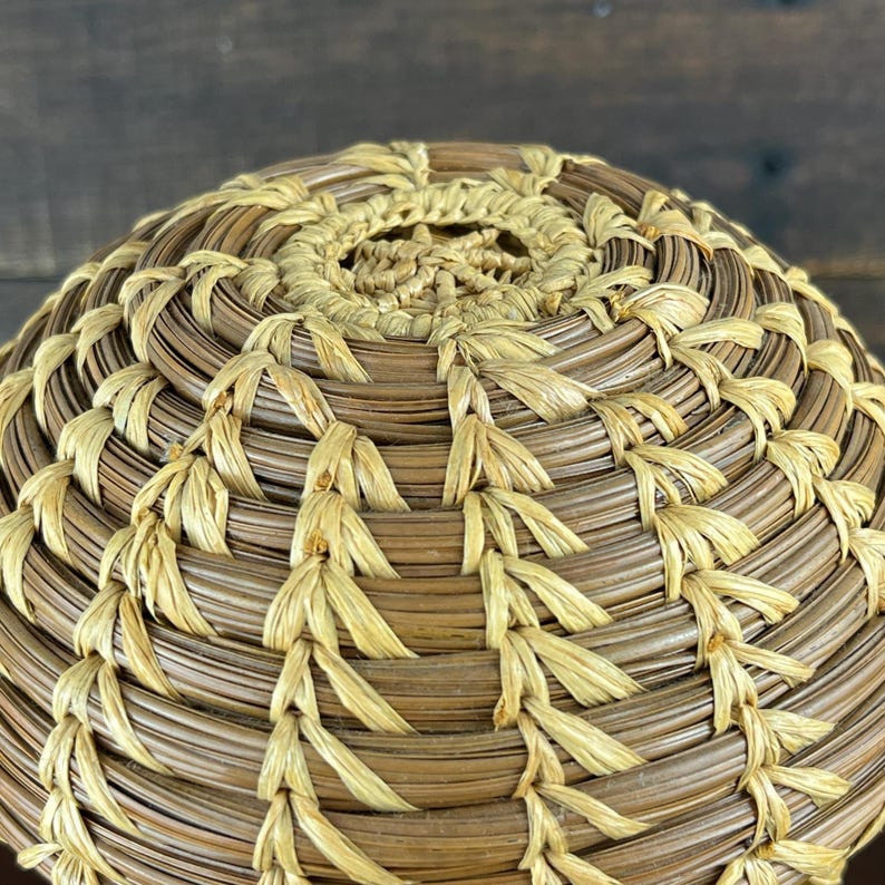 May include: A round, hand-coiled basket lid featuring a dense, circular weave pattern. The construction utilizes alternating rows of dark brown, dried pine needles and light tan, fibrous stitching material, creating a textured, radial design.
