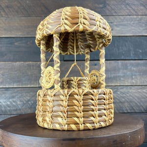 May include: A decorative wishing well crafted from coiled pine needles and light-colored raffia. The structure features a circular base, four vertical support posts, a domed roof, and a small central bucket suspended by a thin fiber cord.