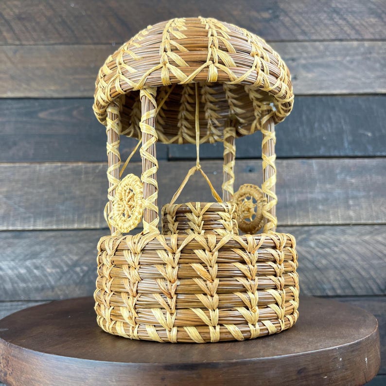 May include: A decorative wishing well crafted from coiled pine needles and light-colored raffia. The structure features a circular base, four vertical support posts, a domed roof, and a small central bucket suspended by a thin fiber cord.