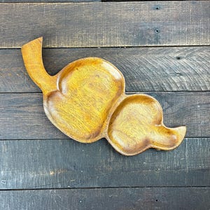 May include: A wooden serving tray with an unusual, organic shape. The tray has three connected sections and a curved handle. The wood is a warm, golden-brown colour with darker brown edges, set against a dark wood background.