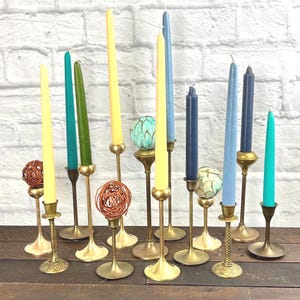 May include: A selection of vintage candlesticks with colourful taper candles. The candles are in shades of yellow, blue, green, and teal. The candle holders are brass and have various designs, including a twisted stem and decorative bases.