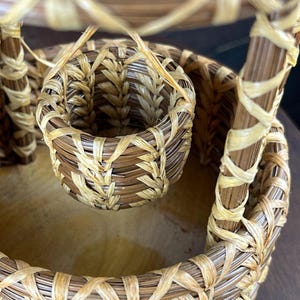 May include: A woven basket structure featuring a small, cylindrical hanging basket suspended by thin, light-colored fibers within a larger, circular base. The object is constructed from dark brown and light tan natural reed or grass materials.