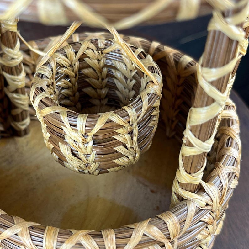 May include: A woven basket structure featuring a small, cylindrical hanging basket suspended by thin, light-colored fibers within a larger, circular base. The object is constructed from dark brown and light tan natural reed or grass materials.