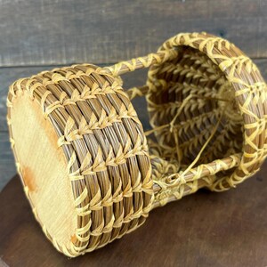 May include: A cylindrical basket crafted from light brown pine needles, featuring a flat wooden base and a woven open-work structure. The basket has a distinct coiled design with visible stitching and a hollow interior, showcasing natural textures.