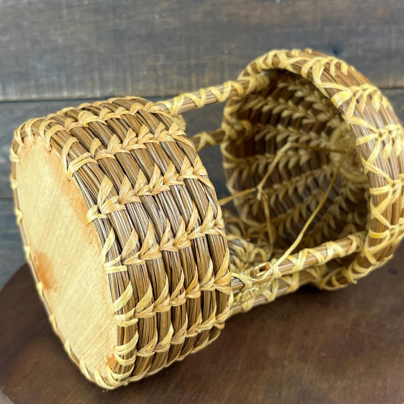 May include: A cylindrical basket crafted from light brown pine needles, featuring a flat wooden base and a woven open-work structure. The basket has a distinct coiled design with visible stitching and a hollow interior, showcasing natural textures.