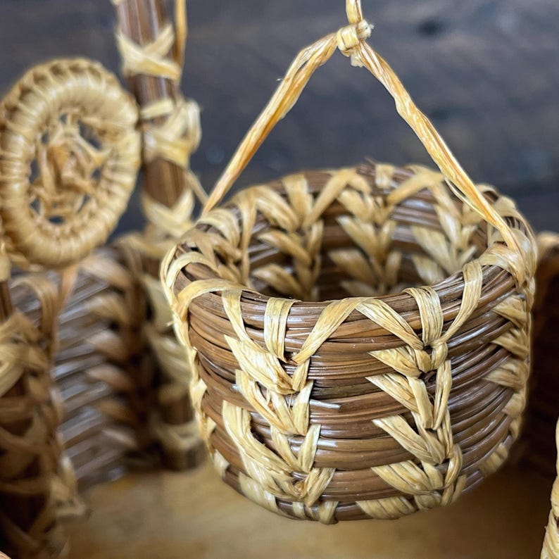 May include: A small, round, woven basket featuring a two-tone pattern of light tan and dark brown natural fibers. The basket has a braided rim and a single handle made of thin, light-colored strips, suspended above a larger woven structure.