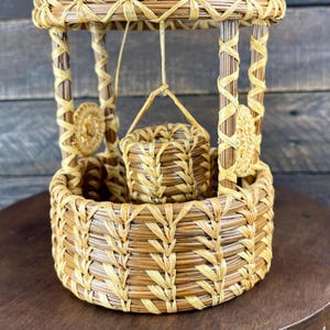 May include: A decorative wishing well crafted from coiled pine needles and light tan raffia. The structure features a circular base, four vertical support posts, a roof, and a small bucket suspended by a thin cord from the center of the roof.