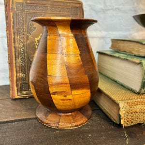 May include: A handcrafted wooden vase with a unique patchwork design. Alternating vertical panels of light and dark brown wood create a striking visual contrast. The vase has a wide base and a flared top, showcasing a blend of natural tones and textures.
