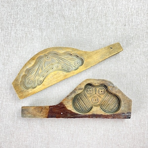 May include: Two antique wooden molds with intricate carved designs. One mold features a floral pattern with circular accents, while the other has a geometric design with circular and linear elements. Both molds have long handles.