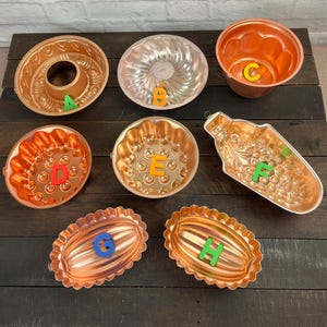 Puede incluir: Una colección de moldes para hornear vintage de color cobre en varias formas, incluyendo diseños de bundt, redondos y ovalados. Los moldes tienen patrones decorativos y están dispuestos sobre una superficie de madera oscura. Cada molde está etiquetado con una letra colorida.