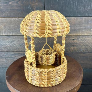 May include: A light brown woven wicker wishing well ornament featuring a domed roof, four vertical support posts, and a small central bucket suspended by a woven cord. The structure is crafted from natural dried plant fibers with a textured finish.