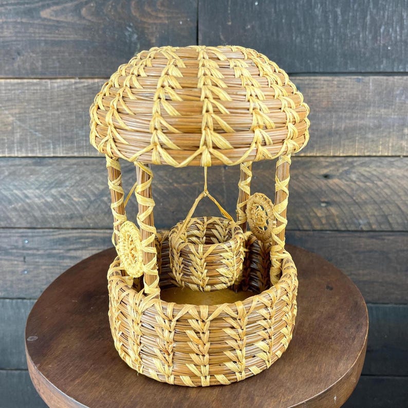 May include: A light brown woven wicker wishing well ornament featuring a domed roof, four vertical support posts, and a small central bucket suspended by a woven cord. The structure is crafted from natural dried plant fibers with a textured finish.