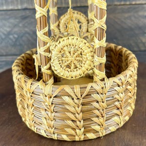 May include: A round, woven basket crafted from light brown pine needles and lighter raffia-like fibers. The basket features a circular base with vertical supports and a decorative, circular woven medallion centered on the front exterior.