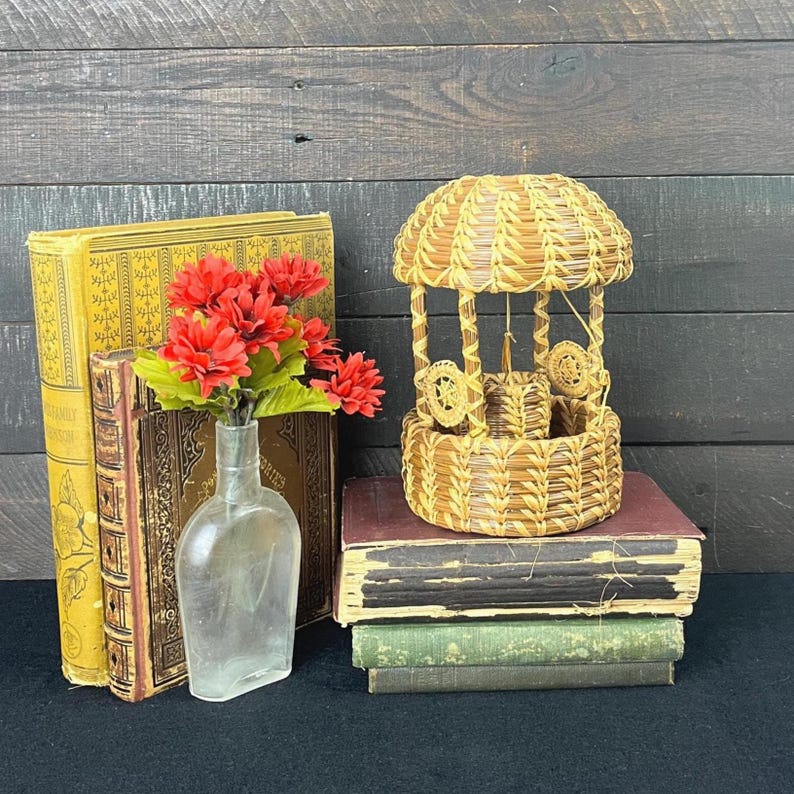 May include: A woven straw wishing well ornament with a domed roof, positioned atop a stack of two vintage books. Beside the books, a clear glass bottle holds a bouquet of artificial red flowers with green leaves, creating a decorative display.