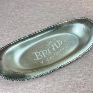 May include: An oval, silver-toned bread tray with a decorative beaded edge. The word "BREAD" is etched in the center of the tray, surrounded by floral accents. The tray is slightly tarnished, giving it a vintage appearance.