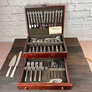 Può includere: Un cofanetto in legno a due livelli contenente un set di posate in acciaio inossidabile Oneida. La sezione superiore contiene forchette e cucchiai, mentre il cassetto inferiore conserva altre posate. Una paletta per dolci e un coltello sono posati accanto.