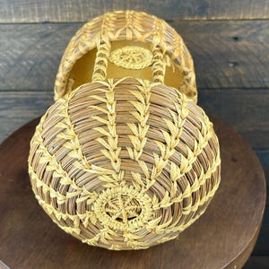 May include: A round, woven basket featuring a light tan and golden yellow straw pattern. The basket has a circular base with a central star-like weave, a domed top section, and an open structure with vertical supports connecting the two parts.