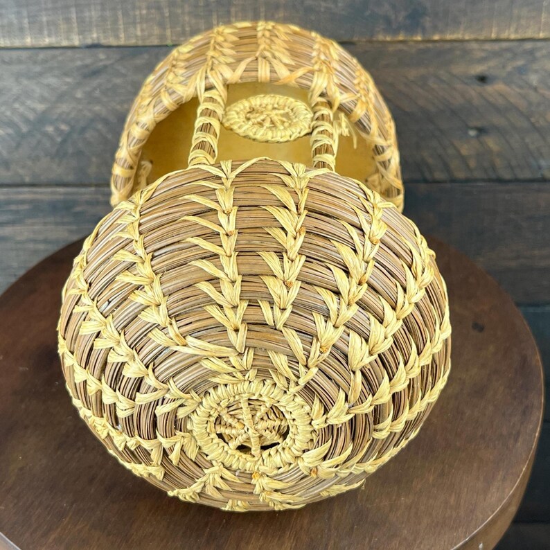 May include: A round, woven basket featuring a light tan and golden yellow straw pattern. The basket has a circular base with a central star-like weave, a domed top section, and an open structure with vertical supports connecting the two parts.