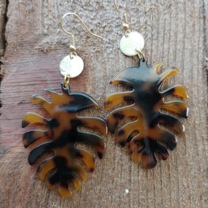 May include: A pair of gold and brown leaf-shaped earrings. The earrings are made of a tortoiseshell-like material and have a gold disc charm hanging from them.