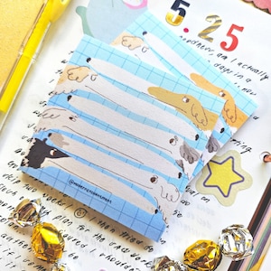 Peut inclure: Une pile de notes autocollantes sur le thème des chiens avec un motif de grille bleu clair. Les notes présentent des illustrations de teckels de différentes couleurs. L'image comprend également un stylo jaune, des bonbons emballés et un journal.