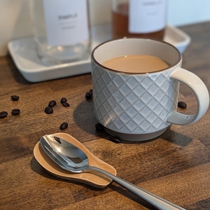 May include: A white ceramic coffee mug with a diamond pattern and a brown rim, filled with coffee, on a wooden surface with a spoon rest and a silver spoon.