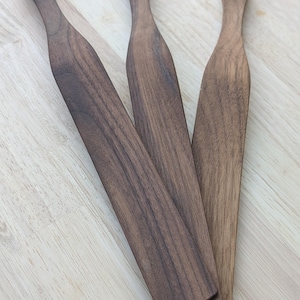Peut inclure: Trois spatules en bois avec une finition marron foncé sont disposées sur une surface en bois clair. Chaque spatule a une longue lame plate et un manche arrondi. Le grain du bois est visible.