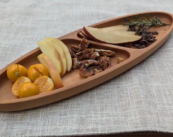 May include: A wooden serving platter with three compartments, containing sliced apples, pecans, yellow cherry tomatoes, pear slices, and chocolate shavings. The oval platter is made of light brown wood.