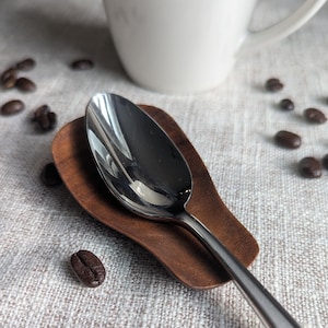 May include: A stainless steel spoon rests on a small, dark brown wooden spoon rest. Coffee beans are scattered around the spoon and rest. A white mug is in the background. The scene is set on a neutral-colored linen surface.