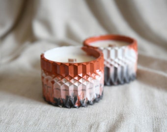 May include: Two cylindrical candles in textured containers featuring a geometric diamond pattern. The containers transition from a terracotta orange at the top to a gradient of white and black at the base, with a visible wooden wick in each candle.