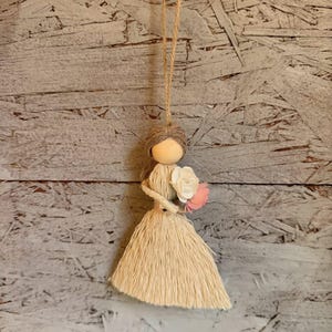 Puede incluir: Adorno de muñeca hecho a mano con cabeza de madera, pelo de hilo y un vestido de hilo color crema. La muñeca sostiene un pequeño ramo de flores de papel blancas y rosas y cuelga de una cuerda de hilo natural.