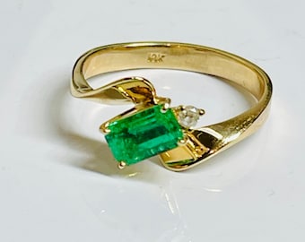 May include: A gold ring with a green emerald gemstone and a small diamond accent. The ring is set in a twisted band.
