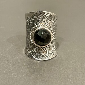 May include: A wide silver ring with an engraved geometric pattern and a large, faceted black gemstone. The ring has a textured surface and a detailed design around the central stone, showcasing a blend of craftsmanship and style.