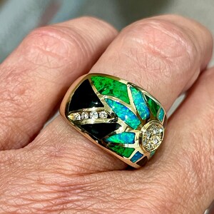 May include: A gold ring with a black, green, and blue opal design. The ring has a large round diamond in the center.