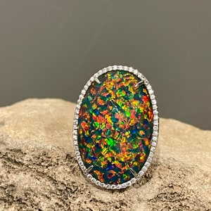 May include: A ring with a large oval-shaped opal set in a white gold band. The opal is a vibrant mix of green, red, and blue colors. The band is accented with a row of small diamonds.