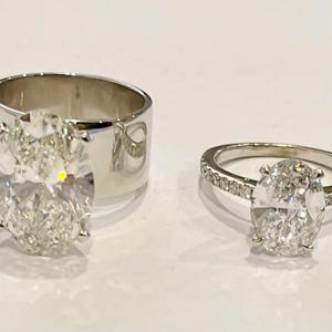May include: Two silver rings with oval-shaped diamonds. The ring on the left has a larger diamond and a wider band. The ring on the right has a smaller diamond and a thinner band.