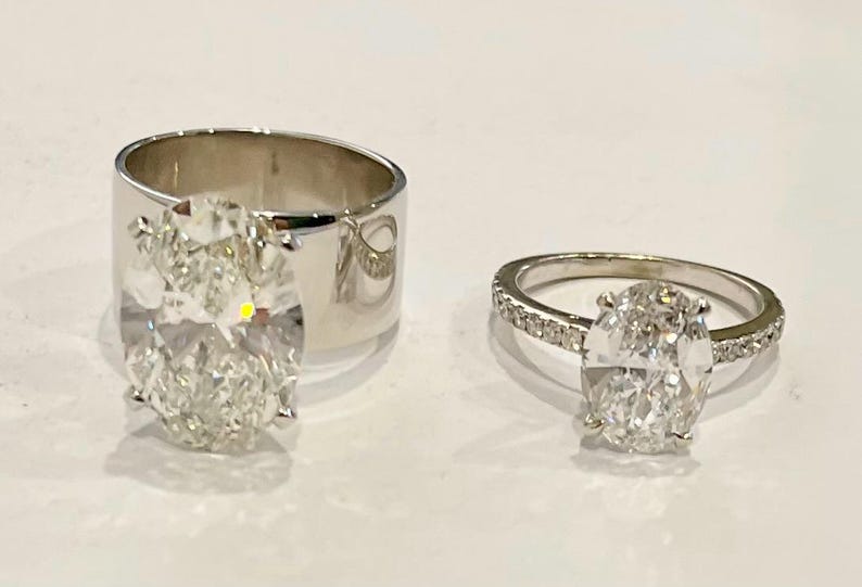 May include: Two silver rings with oval-shaped diamonds. The ring on the left has a larger diamond and a wider band. The ring on the right has a smaller diamond and a thinner band.